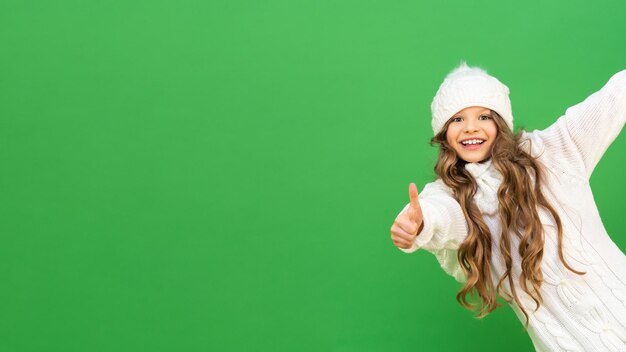 Um lindo modelo com cabelos cacheados em uma camisola branca de inverno em um fundo verde isolado. O bebê olha do canto e mostra um gesto com a mão que está tudo bem.