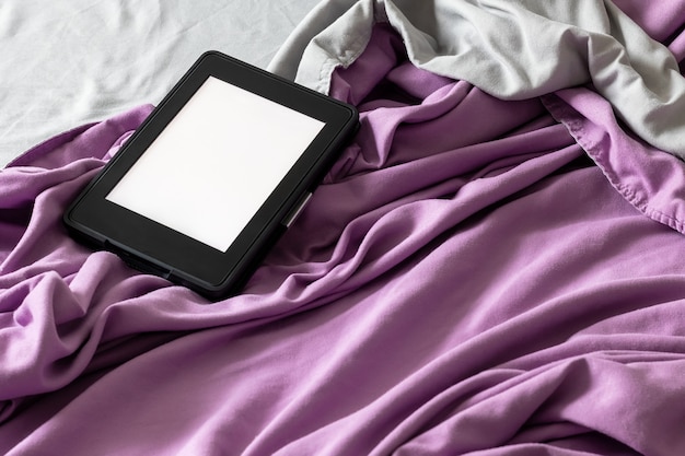 Um livro eletrônico e-reader moderno preto com uma tela em branco em uma cama cinza e roxa.