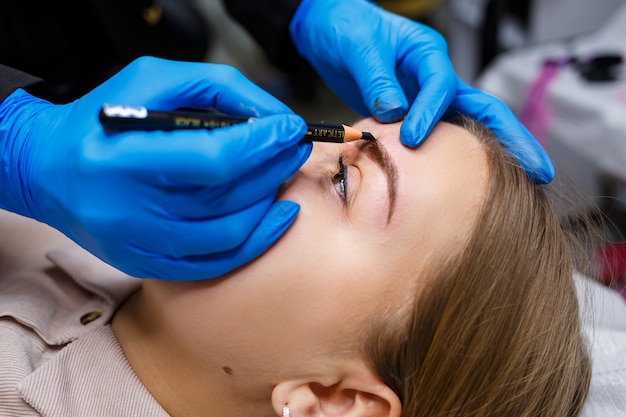 Um maquiador aplica um esboço nas sobrancelhas de uma jovem com um lápis de sobrancelha. Maquilhagem profissional e cuidados cosméticos com a pele.