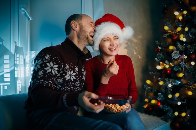 Um marido e sua esposa comem pipoca na véspera de Natal enquanto assistem TV e riem alto