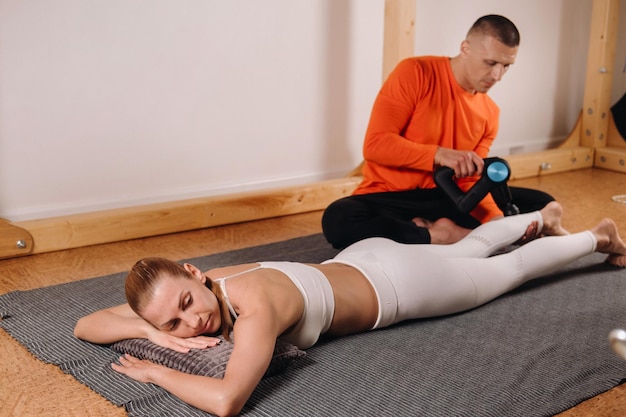 Um massagista masculino massageia o corpo de uma garota com um massageador de percussão para se preparar para um simulador de alongamento
