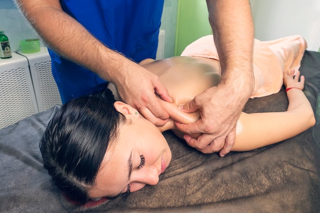 Um massagista profissional faz uma massagem nos ombros de uma jovem. Mulher bonita descansando na cama do spa