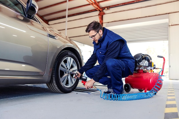 Um mecânico de automóveis no posto de gasolina tenta encher os pneus do carro com a ajuda de um compressor. Um homem em um terno de trabalho azul agachado ao lado de um carro em uma oficina e trabalha com pneus de carros