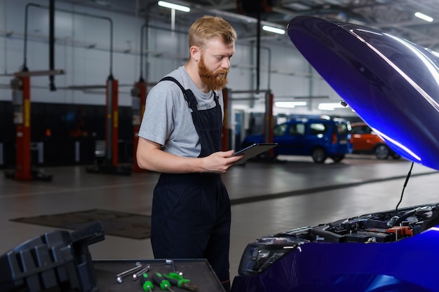 Um mecânico masculino de barba ruiva de uniforme fica em frente ao capô de um carro com um tablet para diagnóstico de erros Estação de diagnóstico profissional