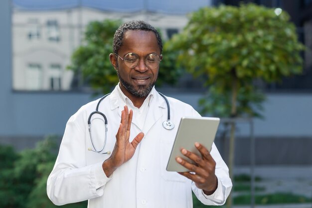 Um médico africano-americano está parado perto do hospital segurando um comprimido nas mãos e fala com