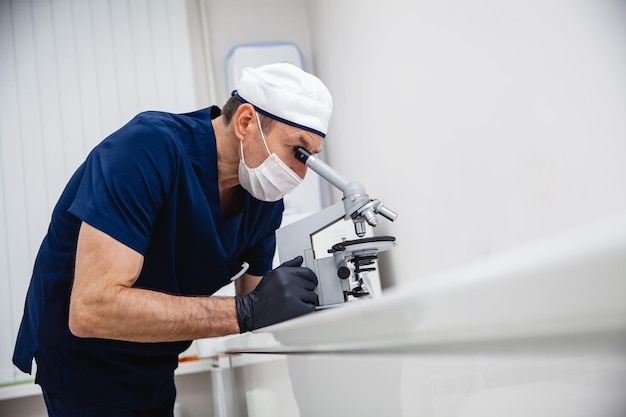 Um médico de terno azul, máscara de chapéu branco e luvas pretas estuda uma lâmina de histologia manchada com um microscópio no laboratório