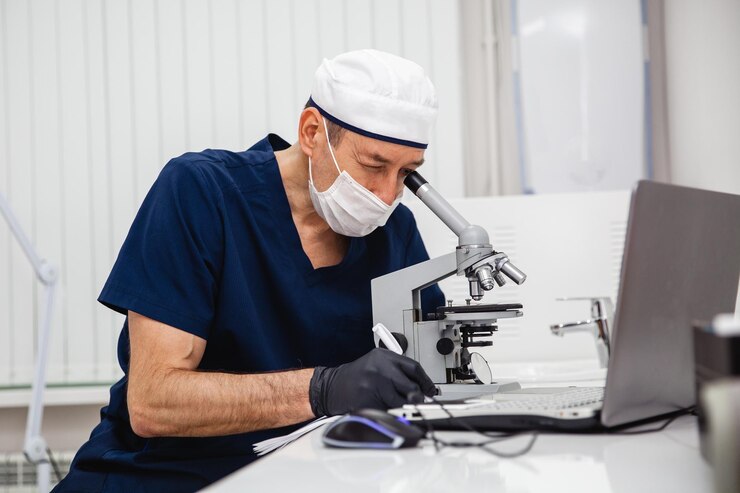 Um médico de terno azul, máscara de chapéu branco e luvas pretas estuda uma lâmina de histologia manchada com um microscópio no laboratório