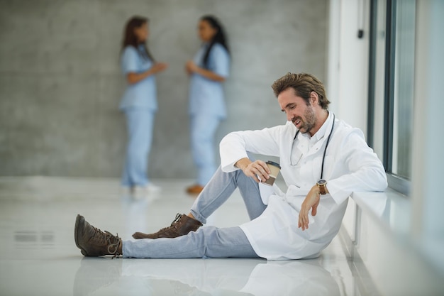 Um médico maduro e estressado sentado no chão perto de uma janela enquanto toma um café rápido em um corredor de hospital durante a pandemia de covid-19.