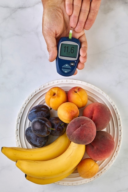 Um medidor de glicose no sangue nas mãos de um homem e um prato de frutas em uma mesa de luz