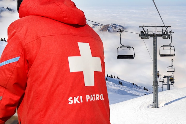 Um membro da patrulha de esqui com uma jaqueta vermelha e uma cruz branca nas costas. Teleférico no topo da montanha