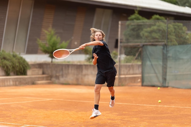 Um menino adolescente de camisa preta jogando tênis no quadro da quadra em movimento batendo de volta a bola