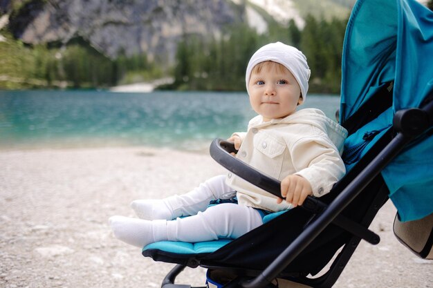 Um menino adorável sentado em um carrinho de bebê perto de um lago de montanha em um dia ensolarado