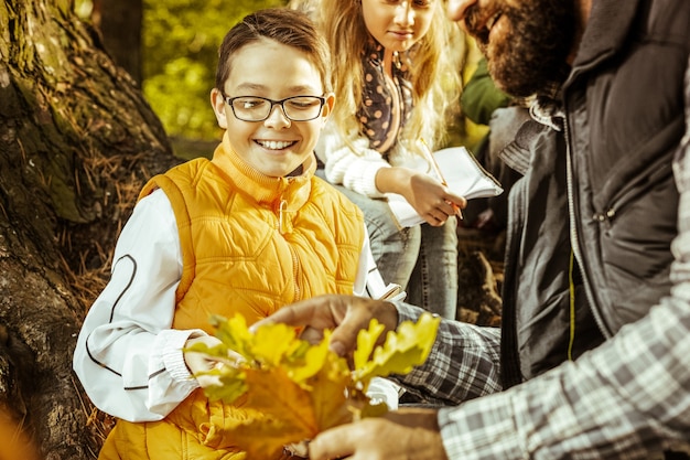 Um menino alegre de óculos e colete laranja olhando para as folhas seguradas por sua professora em um dia bom