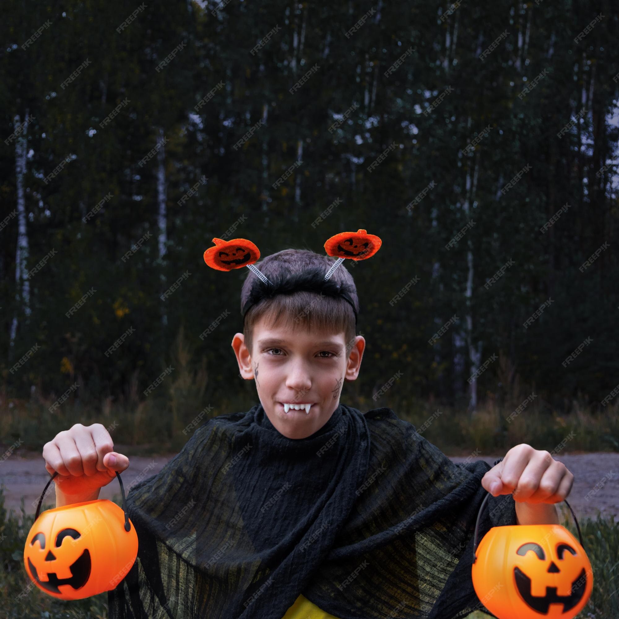 Um menino alegre em fantasias de vampiro mostra seus dentes segurando as  lanternas de jack em suas mãos em um fundo escuro no parque uma criança em  uma fantasia de halloween uma