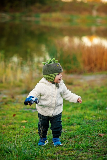 Um menino alegre em um chapéu de malha e roupas quentes, brincando com um carro de brinquedo em uma grama verde perto do lago. Conceito de infância feliz.