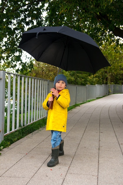 Um menino andando sob um guarda-chuva com uma capa de chuva amarela. Tempo nublado