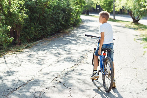 Um menino atencioso senta-se em uma bicicleta azul em um parque sereno, desfrutando do tempo quente em meio à natureza.