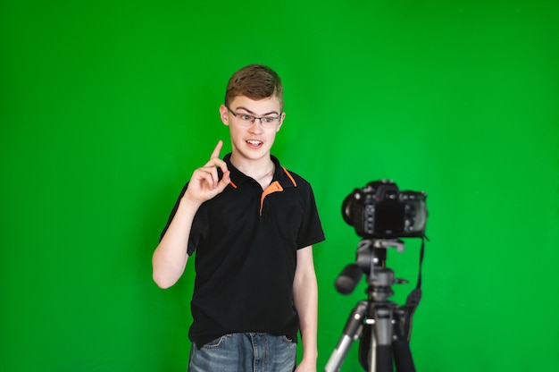 Um menino blogueiro adolescente grava um vídeo para uma câmera contra um fundo verde. Um adolescente caucasiano em um ...