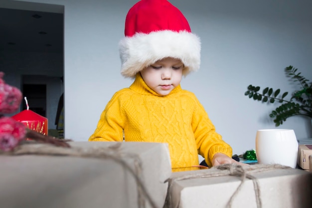 Um menino bonitinho com um suéter amarelo e um chapéu vermelho de Papai Noel em uma mesa de madeira com presentes embalados em papel kraft