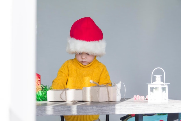 Um menino bonitinho com um suéter amarelo e um chapéu vermelho de Papai Noel em uma mesa de madeira com presentes embalados em papel kraft