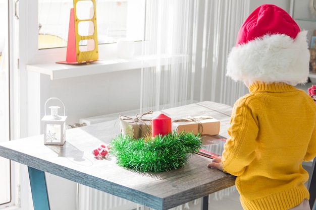 Um menino bonitinho com um suéter amarelo e um chapéu vermelho de Papai Noel em uma mesa de madeira com presentes embalados em papel kraft