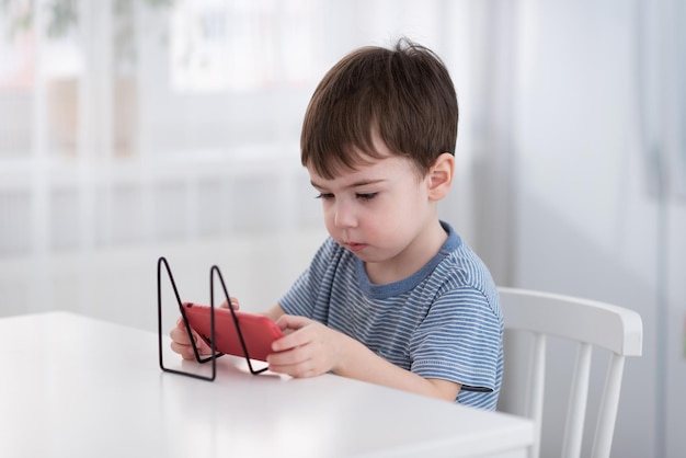 Um menino bonitinho está sentado à mesa e jogando no telefone Conceito de gadgets e crianças