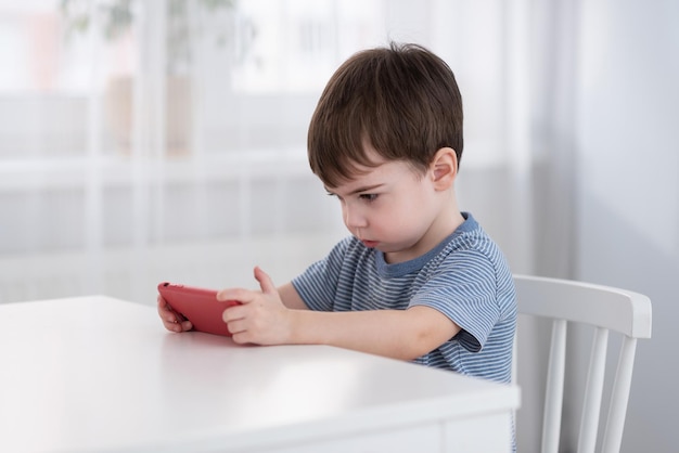 Um menino bonitinho está sentado à mesa e jogando no telefone Conceito de gadgets e crianças