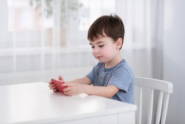 Um menino bonitinho está sentado à mesa e jogando no telefone conceito de gadgets e crianças