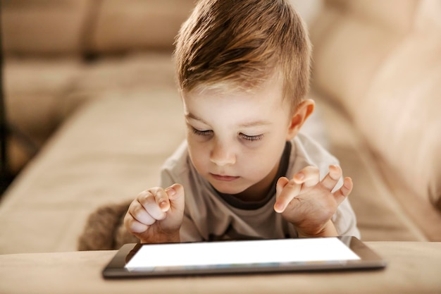 Um menino bonitinho no sofá está jogando no tablet