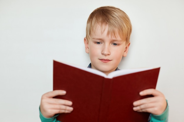 Um menino bonito, com cabelos loiros e olhos azuis, segurando o livro aberto vermelho, lendo histórias interessantes.