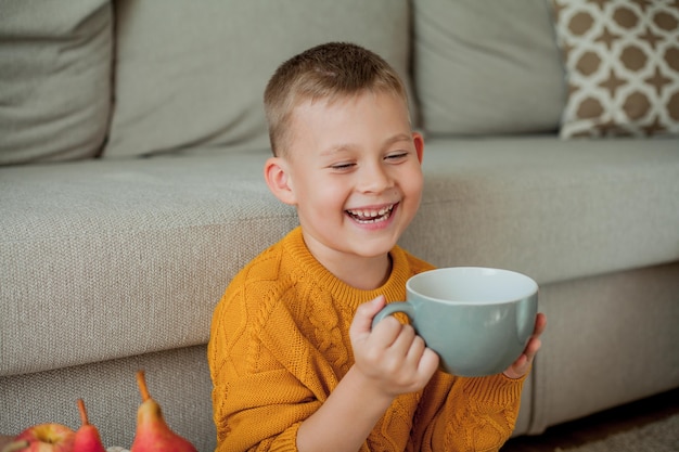 Um menino bonito com um suéter laranja está bebendo chá. Aconchegante retrato de um menino sentado em casa. Outono.