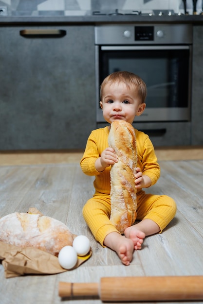 Um menino bonito de um ano está sentado na cozinha e comendo um pão comprido ou baguete na cozinha A primeira vez que uma criança come pão O pão é bom para as crianças