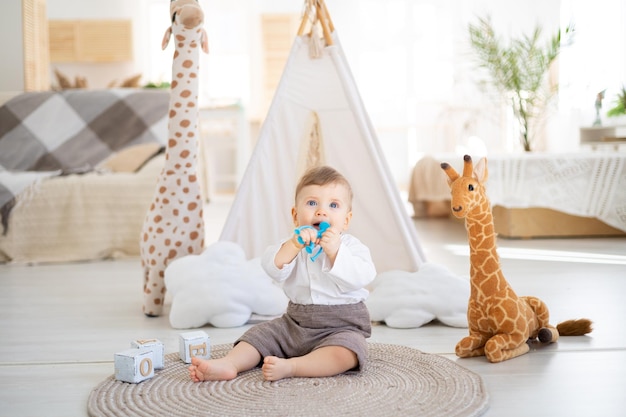 Um menino bonito e saudável está sentado em um tapete na luminosa sala de estar da casa no contexto de uma tenda e brinquedos de pelúcia brincando com brinquedos educativos de madeira têxteis domésticos