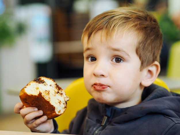 Foto um menino bonito em roupas casuais a comer um bolo de chocolate numa praça de refeições.