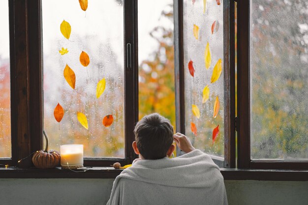 Um menino bonito embrulhado em um cobertor bebe chá quente e olha pela janela aberta para a maravilhosa natureza do outono Decoração da casa do outono Humor de outono aconchegante Ação de Graças Halloween
