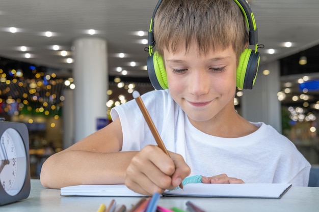 Um menino bonito ouve música com fones de ouvido e escreve lição de casa em um caderno em um café bokeh closeup tecnologia e música na vida das crianças lição escolar remota on-line