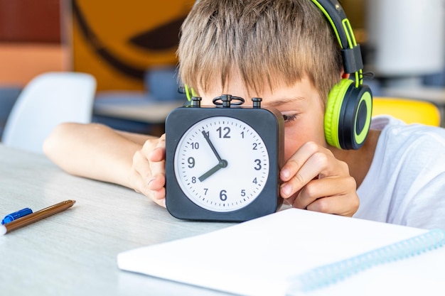Um menino bonito ouve música com fones de ouvido mantém um despertador em um café bokeh closeup Hora de ouvir música