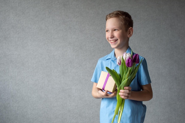 Um menino bonito ri e segura um buquê colorido de tulipas e uma caixa com um presente nas mãos
