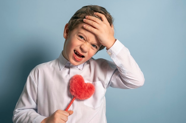 Um menino bonito segura um coração vermelho fofo O menino segura a cabeça e grita algo irritado