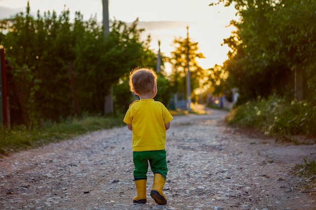 Um menino caminha pela estrada rural com botas de borracha amarela e uma camiseta brilhante. li