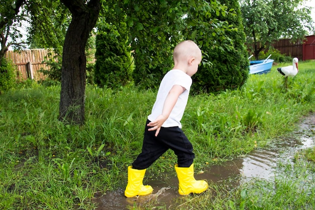 Um menino careca de botas amarelas corre no campo através de poças ao ar livre