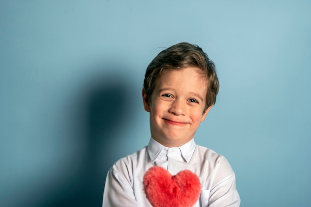 Um menino caucasiano bonito em uma camisa branca tem um coração vermelho fofo em suas mãos órfãos do Dia das Mães