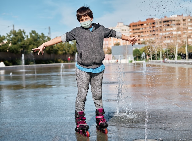 Um menino caucasiano com uma máscara e patins, patinando na água na fonte do parque. Nova pandemia normal Covid 19