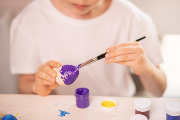 Um menino caucasiano em uma camiseta branca pinta ovos para celebrar o feriado da Páscoa com cores brilhantes