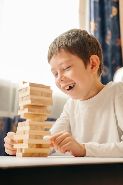 Um menino caucasiano sorridente brinca em casa, na mesa de Jenga. Jogos de tabuleiro para crianças e adultos. Um passatempo sem gadgets. Jogos durante as férias em casa. Criança feliz brinca com blocos de madeira.