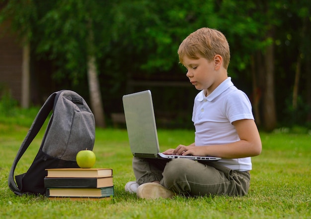 Um menino caucasiano sorridente com uma maleta senta-se no gramado e lê um livro o conceito de educação
