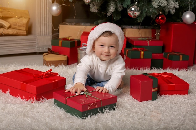 Um menino com chapéu de papai noel está sentado perto da lareira e árvores de natal ao lado de caixas de presente