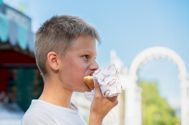 Um menino com fome saboreando um cachorro-quente ao ar livre em um dia ensolarado tomando uma mordida satisfatória em um parque público