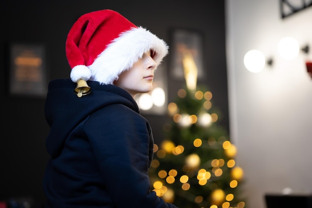 Um menino com o chapéu de pele de Natal do Papai Noel está sentado de lado e observa as luzes brilhantes do Natal