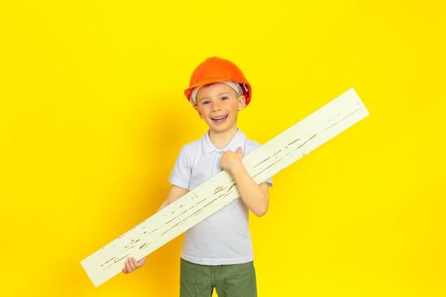 Um menino com um capacete de construção laranja em uma parede amarela segurando uma placa de madeira nas mãos, ele está feliz e sorridente.